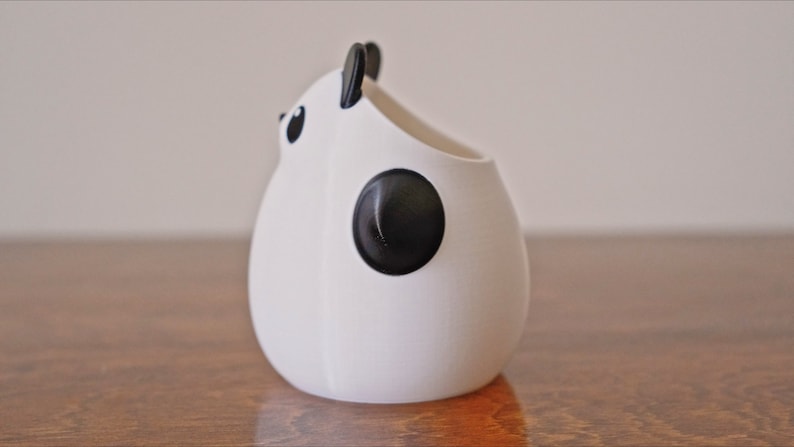 Op de afbeelding: Een witte keramische panda-vormige plantenpot met zwarte oren, ogen en een vlek op zijn rug. De pot staat op een houten oppervlak.