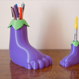 May include: Two purple eggplant-shaped pencil holders with green tops. The larger holder has multiple pens and markers inside. The smaller holder has one yellow marker inside.