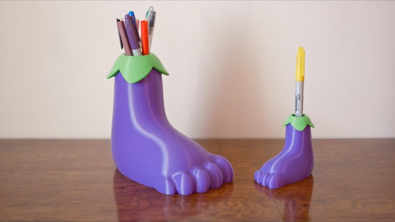 May include: Two purple eggplant-shaped pencil holders with green tops. The larger holder has multiple pens and markers inside. The smaller holder has one yellow marker inside.