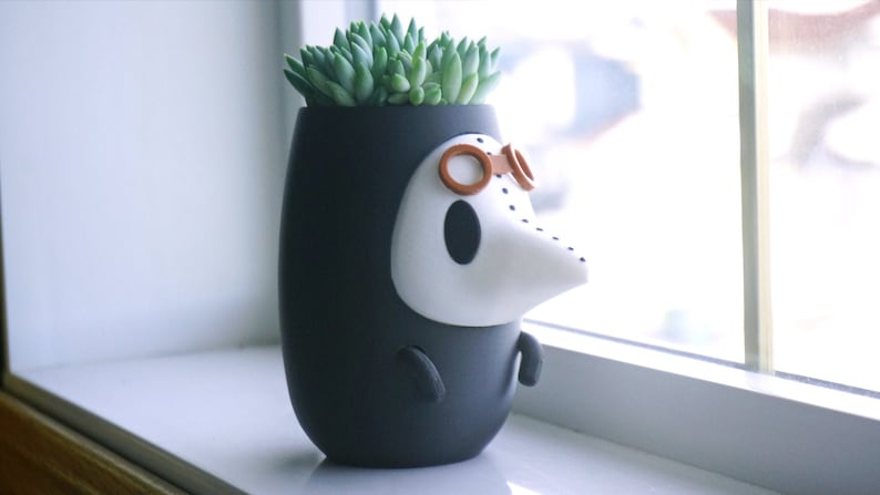May include: A black and white ceramic planter shaped like a character wearing goggles. The planter has a white face with a long nose and is holding a small green succulent plant.