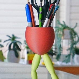 May include: A red frog-shaped desk organizer with green legs holds a variety of writing utensils, including pens, pencils, and scissors.