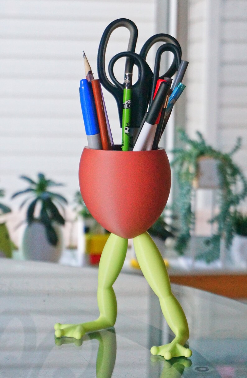 May include: A red frog-shaped desk organizer with green legs holds a variety of writing utensils, including pens, pencils, and scissors.
