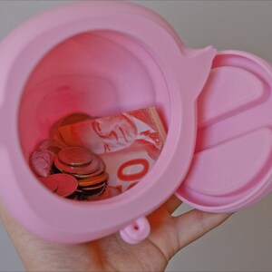 May include: A pink silicone piggy bank with a removable lid. The piggy bank is open and contains coins and a Canadian five dollar bill.