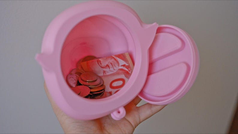 May include: A pink silicone piggy bank with a removable lid. The piggy bank is open and contains coins and a Canadian five dollar bill.