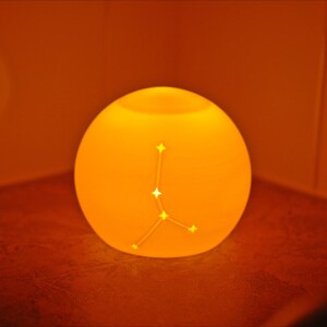 May include: A glowing, spherical lamp with a constellation design. The lamp emits a warm orange light, creating a cozy ambiance. The constellation is a series of connected stars. The lamp is placed in a corner.