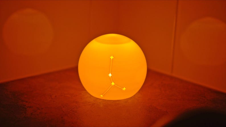 May include: A glowing, spherical lamp with a constellation design. The lamp emits a warm orange light, creating a cozy ambiance. The constellation is a series of connected stars. The lamp is placed in a corner.