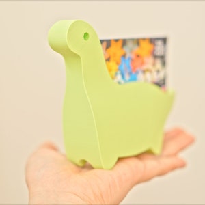 May include: A light green dinosaur-shaped card holder made of plastic. The holder is designed to hold business cards or other small items.