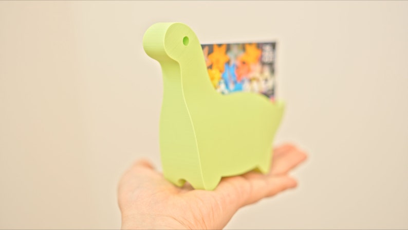 May include: A light green dinosaur-shaped card holder made of plastic. The holder is designed to hold business cards or other small items.
