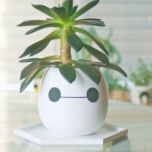 Puede incluir: Maceta de cer&aacute;mica blanca con forma de robot con ojos negros y boca, que contiene una planta suculenta verde. La maceta est&aacute; sobre un platillo hexagonal blanco.