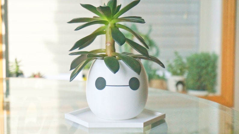 Puede incluir: Maceta de cer&aacute;mica blanca con forma de robot con ojos negros y boca, que contiene una planta suculenta verde. La maceta est&aacute; sobre un platillo hexagonal blanco.