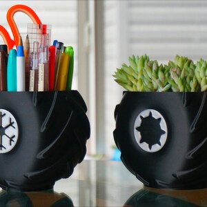 May include: Two black tire-shaped planters with white designs. One planter holds a variety of pens and pencils, while the other holds a small succulent plant.