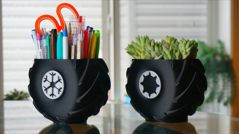 May include: Two black tire-shaped planters with white designs. One planter holds a variety of pens and pencils, while the other holds a small succulent plant.