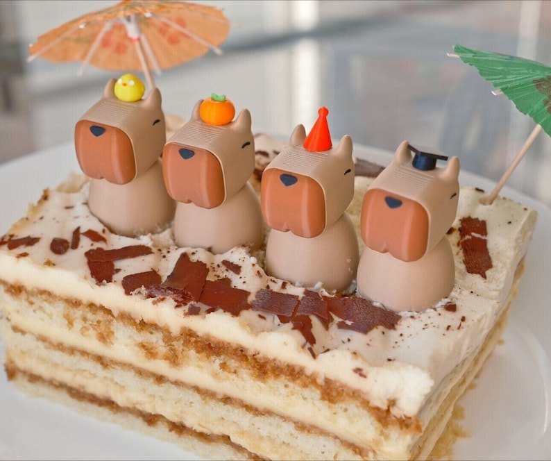 May include: A multi-layered tiramisu cake decorated with four brown and beige animal figurines wearing party hats. The cake is topped with chocolate shavings and a small green umbrella.