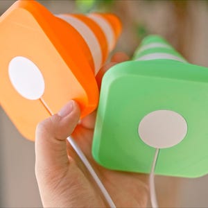 May include: Two colorful, square-shaped objects with cone tops are held in a hand. One is orange with white stripes, the other is green with white stripes. Each has a white circle on the square base and a white cord.