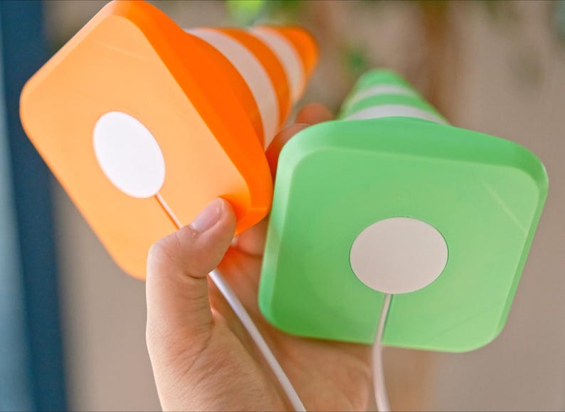 May include: Two colorful, square-shaped objects with cone tops are held in a hand. One is orange with white stripes, the other is green with white stripes. Each has a white circle on the square base and a white cord.