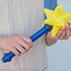 K&ouml;nnte beinhalten: Ein blau-gelber Zauberstab in Sternform. Der Zauberstab hat einen blauen Griff und einen leuchtend gelben Stern an der Spitze. Der Zauberstab wird von einer Person gehalten, die ein blaues Hemd und eine beige Sch&uuml;rze tr&auml;gt.