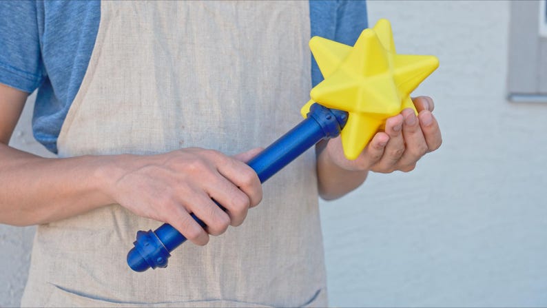 K&ouml;nnte beinhalten: Ein blau-gelber Zauberstab in Sternform. Der Zauberstab hat einen blauen Griff und einen leuchtend gelben Stern an der Spitze. Der Zauberstab wird von einer Person gehalten, die ein blaues Hemd und eine beige Sch&uuml;rze tr&auml;gt.