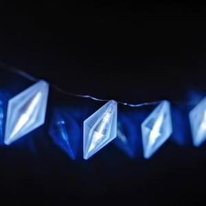 May include: A string of blue diamond-shaped LED lights against a dark background. The lights are strung on a thin wire, creating a festive and decorative element. The lights are a bright, cool blue.