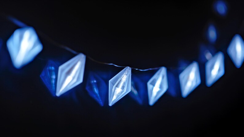 May include: A string of blue diamond-shaped LED lights against a dark background. The lights are strung on a thin wire, creating a festive and decorative element. The lights are a bright, cool blue.