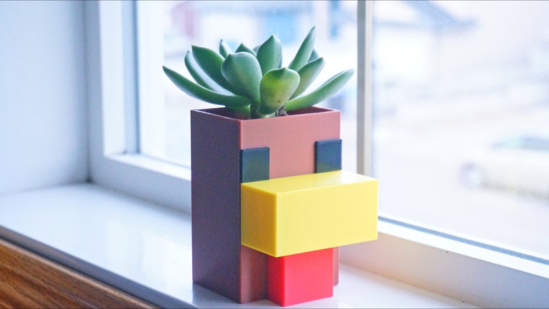 K&ouml;nnte beinhalten: Ein brauner, gelber und roter Pflanzer in Form eines Minecraft-Huhns, mit einer gr&uuml;nen Sukkulente, die darin w&auml;chst.