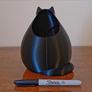 May include: A black cat-shaped desk organizer with a pen next to it. The organizer is 3D-printed and has a hollow interior. The pen is a Sharpie Fine Point marker. The cat has a rounded body and a small tail.
