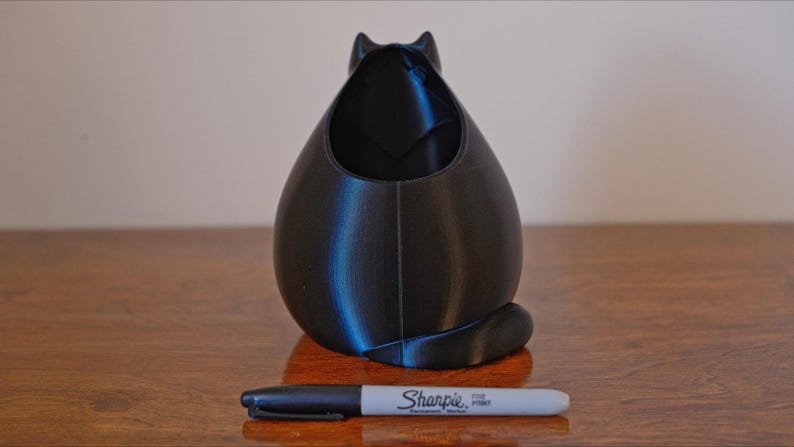 May include: A black cat-shaped desk organizer with a pen next to it. The organizer is 3D-printed and has a hollow interior. The pen is a Sharpie Fine Point marker. The cat has a rounded body and a small tail.