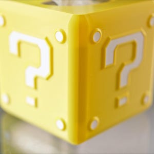 K&ouml;nnte beinhalten: Ein gelber Fragezeichenblock mit wei&szlig;en Fragezeichen auf jeder Seite. Der Block ist ein beliebtes Symbol aus der Super Mario-Videospielserie.
