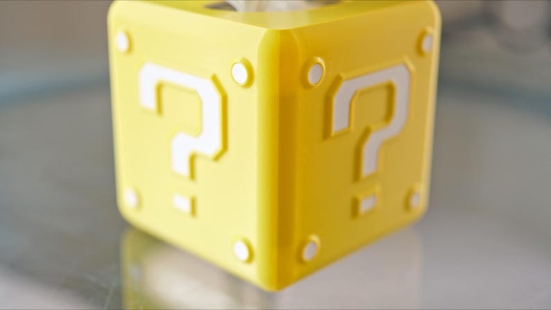 K&ouml;nnte beinhalten: Ein gelber Fragezeichenblock mit wei&szlig;en Fragezeichen auf jeder Seite. Der Block ist ein beliebtes Symbol aus der Super Mario-Videospielserie.
