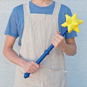 K&ouml;nnte beinhalten: Ein gelber Zauberstab in Sternform mit blauem Griff. Die Person tr&auml;gt ein blaues T-Shirt und eine beige Sch&uuml;rze. Der Zauberstab ist ein verspieltes Spielzeug oder eine Requisite f&uuml;r fantasievolles Spielen oder Kost&uuml;me.