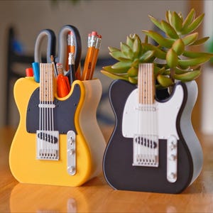 Puede incluir: Dos organizadores de escritorio con forma de guitarra, uno amarillo y otro negro, están uno al lado del otro sobre una superficie de madera. El organizador amarillo contiene bolígrafos y tijeras. El organizador negro contiene una planta suculenta verde. Ambos tienen detalles de guitarra.
