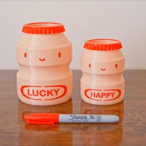 May include: Two pink, cartoon-style bottles with orange lids and smiling faces. The larger bottle has the word "LUCKY" in red, and the smaller bottle says "HAPPY." A red and gray Sharpie marker rests in front of them.
