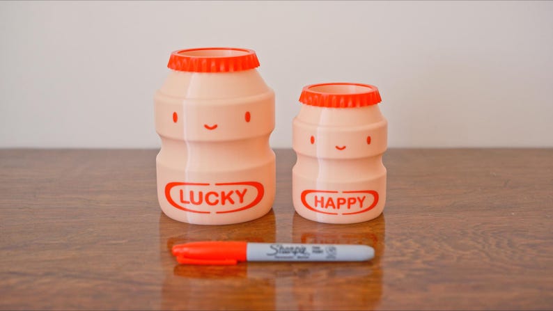 May include: Two pink, cartoon-style bottles with orange lids and smiling faces. The larger bottle has the word "LUCKY" in red, and the smaller bottle says "HAPPY." A red and gray Sharpie marker rests in front of them.