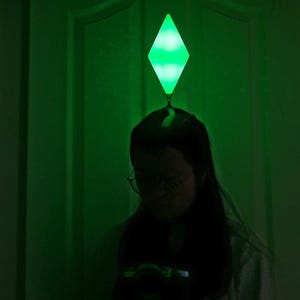 May include: A person is silhouetted in front of a green-lit door, with a glowing green diamond-shaped object above their head. The object resembles the plumbob from The Sims video game series. The scene is dimly lit, with a focus on the glowing object.