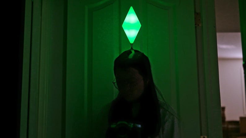 May include: A person is silhouetted in front of a green-lit door, with a glowing green diamond-shaped object above their head. The object resembles the plumbob from The Sims video game series. The scene is dimly lit, with a focus on the glowing object.