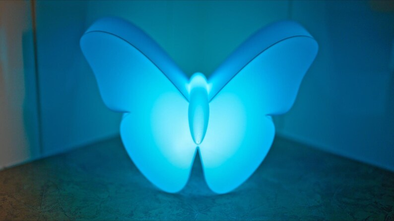 May include: A glowing blue butterfly-shaped lamp. The lamp has a smooth, rounded design with a soft, diffused light. The wings are spread wide, and the body is centered, creating a symmetrical and calming aesthetic. The lamp is a decorative lighting fixture.