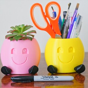 May include: Two 3D printed smiley face planters, one pink with a succulent plant and the other yellow with a variety of pens and scissors.