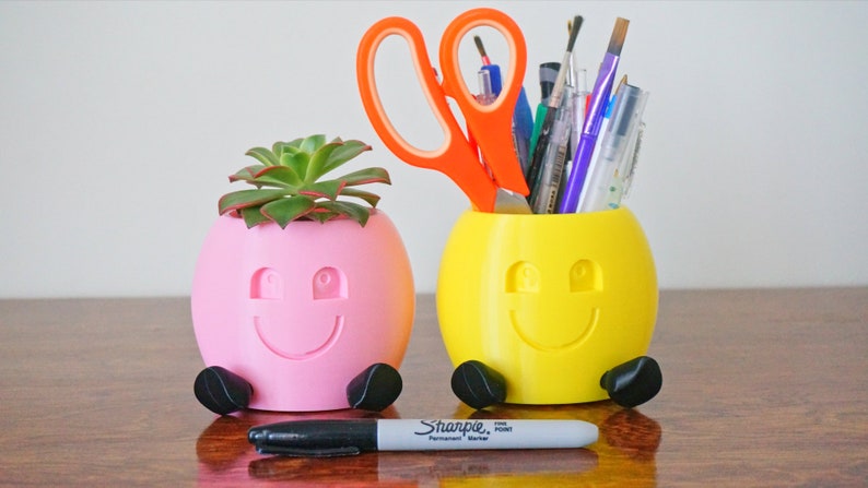 May include: Two 3D printed smiley face planters, one pink with a succulent plant and the other yellow with a variety of pens and scissors.