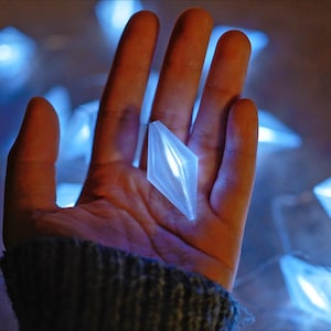 May include: A hand holds a glowing, diamond-shaped light. The light is white and surrounded by a string of similar blue lights. The background is dark and out of focus, creating a soft, ethereal glow.