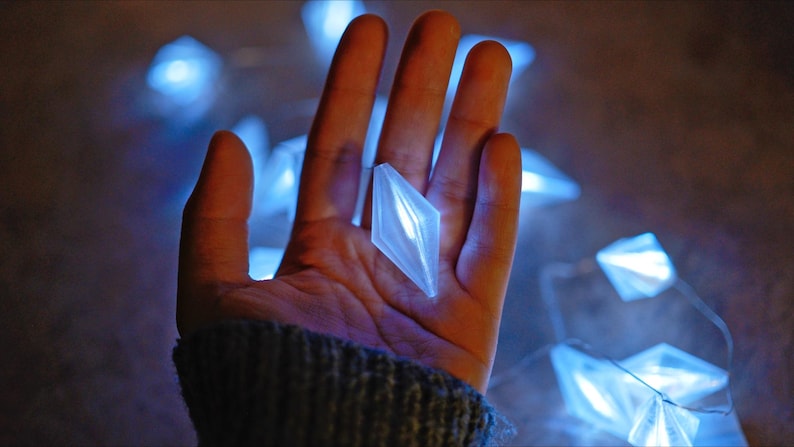 May include: A hand holds a glowing, diamond-shaped light. The light is white and surrounded by a string of similar blue lights. The background is dark and out of focus, creating a soft, ethereal glow.