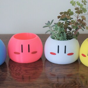 May include: Four colorful ceramic planters shaped like Japanese dango mochi, a sweet rice cake. The planters are blue, pink, white, and yellow. The white planter has a small green succulent plant growing in it.