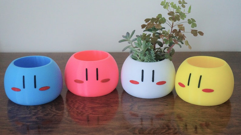 May include: Four colorful ceramic planters shaped like Japanese dango mochi, a sweet rice cake. The planters are blue, pink, white, and yellow. The white planter has a small green succulent plant growing in it.
