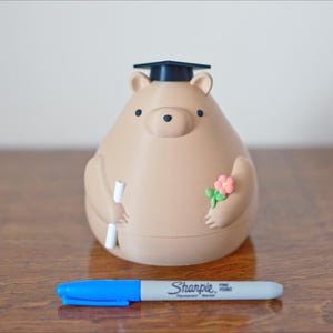 Puede incluir: Una hucha con forma de oso marr&oacute;n claro con birrete, que sostiene un diploma y flores. Un rotulador Sharpie azul y gris est&aacute; delante de la hucha. La hucha est&aacute; sobre una superficie de madera.