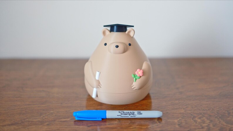 Puede incluir: Una hucha con forma de oso marr&oacute;n claro con birrete, que sostiene un diploma y flores. Un rotulador Sharpie azul y gris est&aacute; delante de la hucha. La hucha est&aacute; sobre una superficie de madera.