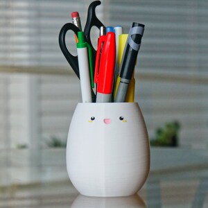 May include: White ceramic pencil holder shaped like an egg with a cute cartoon face. The holder has a yellow mouth and two small yellow eyes. It is filled with various pens, pencils, and a pair of scissors.