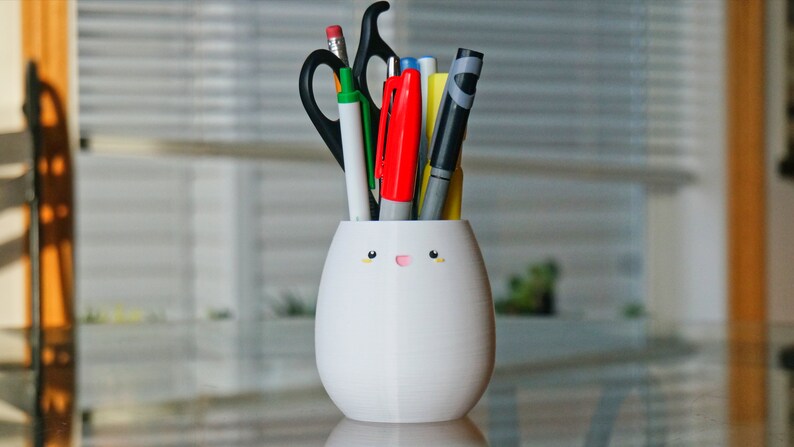 May include: White ceramic pencil holder shaped like an egg with a cute cartoon face. The holder has a yellow mouth and two small yellow eyes. It is filled with various pens, pencils, and a pair of scissors.
