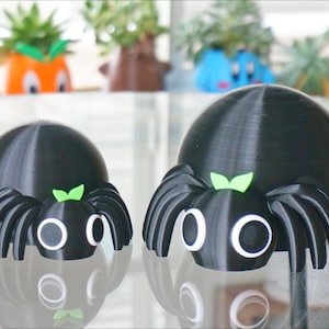 May include: Two black 3D printed spiders with white eyes and green leaves on their heads. The larger spider is in the foreground and the smaller spider is in the background.