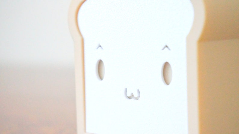 May include: A white bread-shaped object with a cute cartoon face. The face has two small black eyes, a small black mouth, and two small black eyebrows.