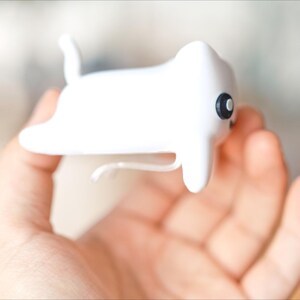 May include: A white, cat-shaped ornament with a black eye and a curved tail. The object has a smooth, rounded form and is held in a hand. The background is blurred, suggesting a shallow depth of field. The object appears to be a decorative item or a small toy.