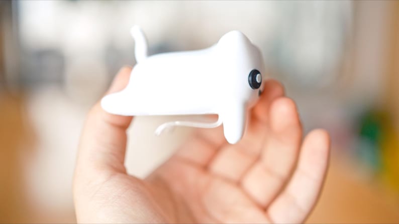May include: A white, cat-shaped ornament with a black eye and a curved tail. The object has a smooth, rounded form and is held in a hand. The background is blurred, suggesting a shallow depth of field. The object appears to be a decorative item or a small toy.