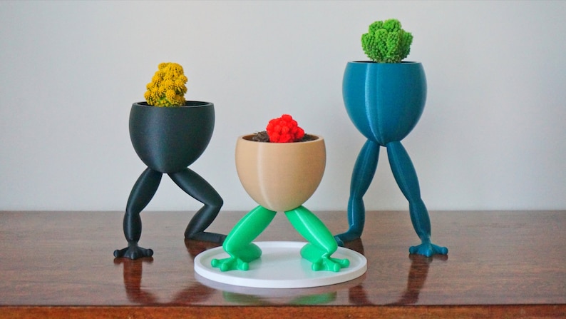 May include: Three frog-leg plant pots with different colored legs and pots. The pots are holding small plants. The pots are on a wooden surface.